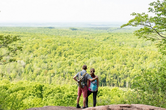 Man and woman looking down and viewing a wide landscape of bring green forests.