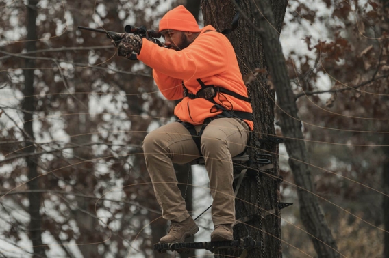Person sitting in tree stand wearing blaze orange and aiming a rifle.