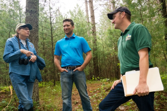 Three people standing in a forest clearing. The one farthest to the right is holding a stack of folders.