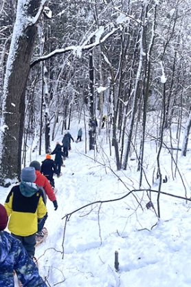 Kids snowshoe in a line through winter woods. 