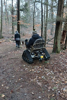 Park visitor driving tracked outdoor wheelchair on a trail through a forest.