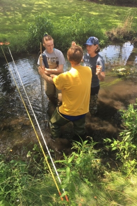 A photo of an instructor teaching two students how to use a flow meter, standing in a shallow stream.