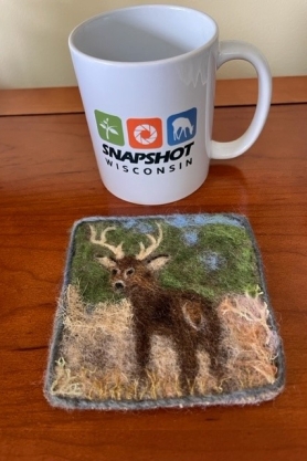 An image of a wool felted coaster and a coffee cup. The wool felted coaster has a buck in grass.