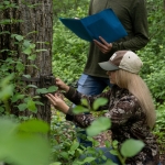 Two young adults check on a trail camera, surrounded by green forest.