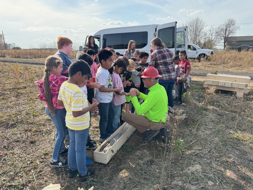 A group of Abbotsford students and teachers standing outside in a field surrounding a worker in a hard hat who is teaching them to collect soil samples.