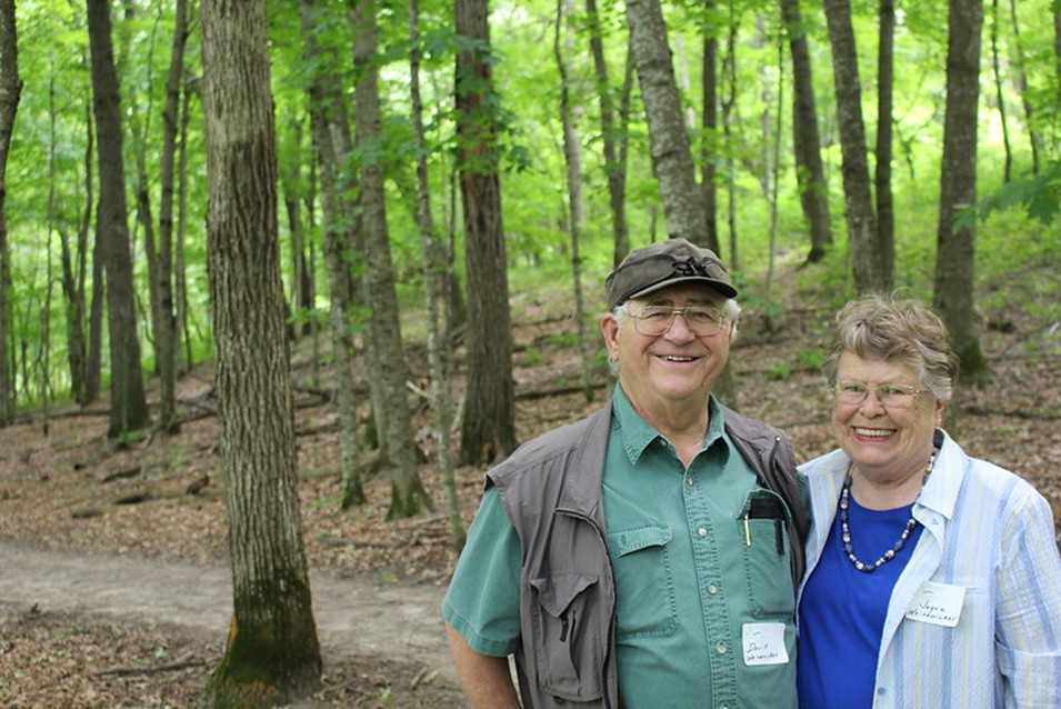 An older couple pictured in front of a trail in the green lush woods.