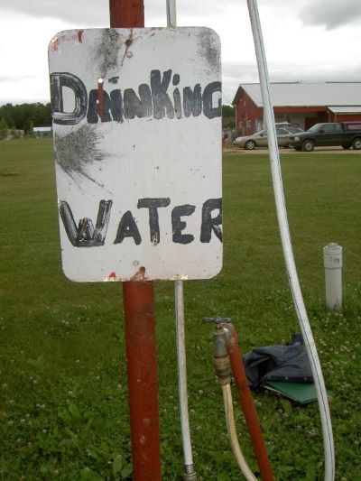 A sign with Drinking Water written on it in marker.  It marks an old well in the background with a hose coming out of it.