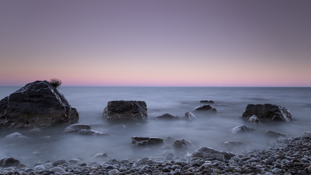 "Belt of Venus" photo by Kevin O'Donnell. 2019 Great Waters Photo Contest entry. The photo shows mist over small boulders on a rocky beach with a pink sunset or sunrise in the distance over the vast water.