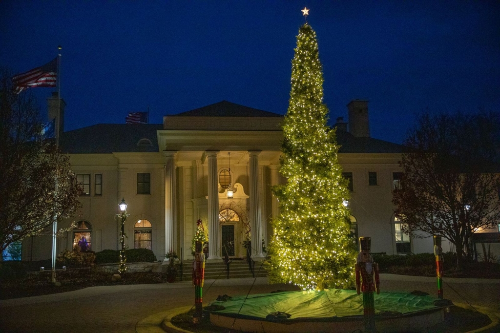 An evergreen decorated with white Christmas lights in front of a very fancy home.