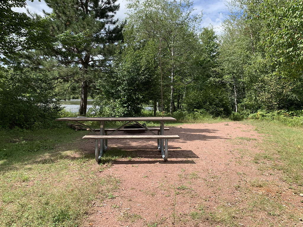 Accessible canoe campsite with a crushed red gravel camping surface, accessible fire ring and accessible picnic table, surrounded by trees along the shoreline of the Turtle-Flambeau Scenic Waters Area.